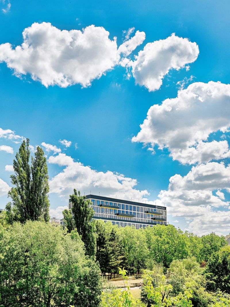 Ein modernes mehrstöckiges Bürogebäude, eingerahmt von hohen grünen Bäumen vor einem leuchtend blauen Himmel mit weißen Wolken