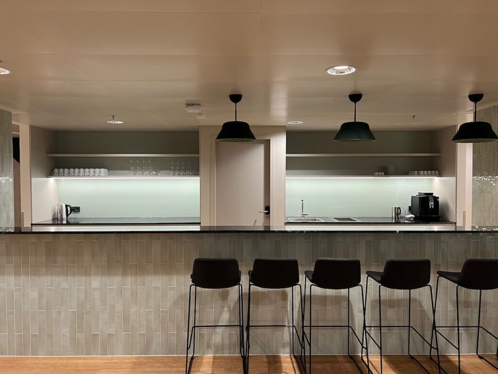 A modern office pantry area featuring a long counter with black bar stools, a tiled backsplash, and open shelving with glassware and coffee machine.