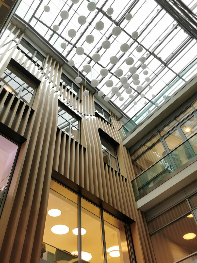 Interior view of a modern building featuring vertical wooden slats, large glass windows, and a glass atrium roof decorated with hanging sphere lights.