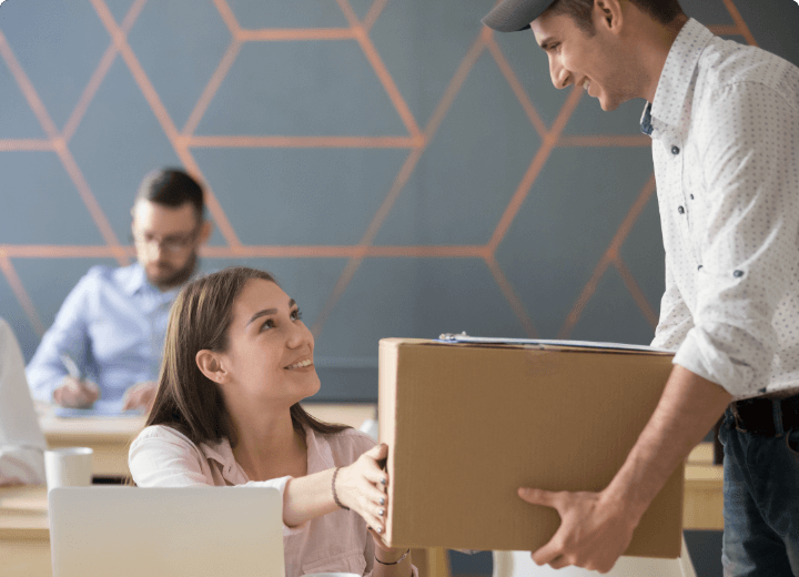 Paketdienst liefert einen Karton an eine Mitarbeiterin an ihrem Schreibtisch im Büro, während Kollegen im Hintergrund arbeiten