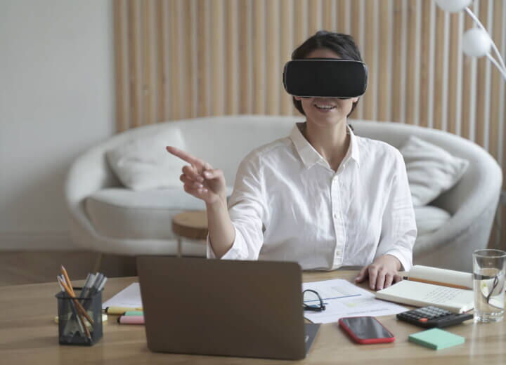 A woman wearing a VR headset and a white shirt, sitting at a desk with a laptop and pointing at a virtual screen in a modern living room.