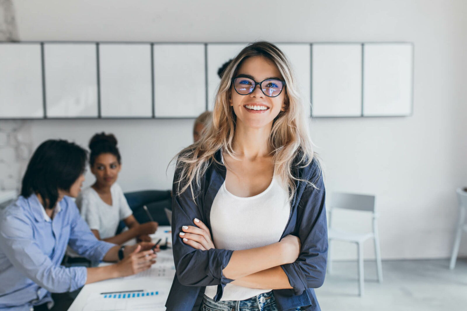Smiling blonde female secretary wearing trendy glasses with arms crossed in a modern office during a meeting