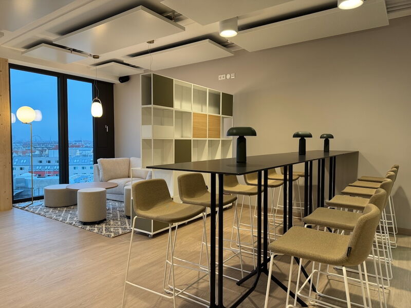 Spacious modern office space featuring a high standing desk with green stools, a cozy lounge area with a sofa, and a large window overlooking a cityscape view at dusk.