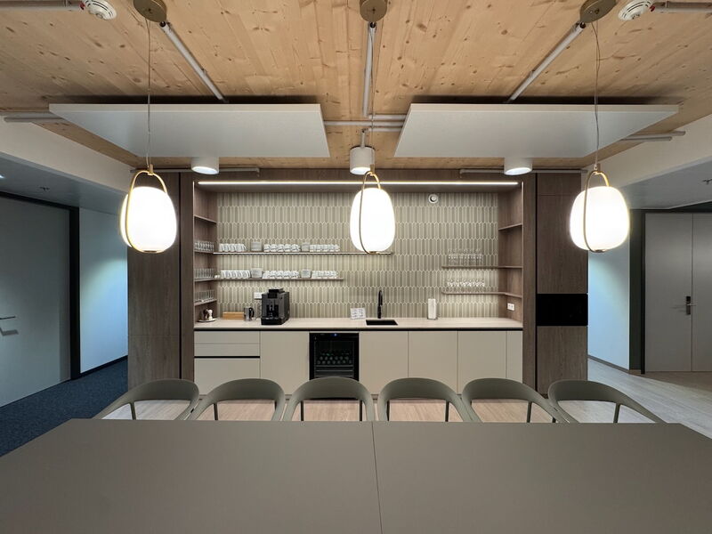 Sleek modern office kitchenette featuring wooden ceiling panels, stylish pendant lighting, organized shelving with mugs and glasses, and a long meeting table in the foreground.