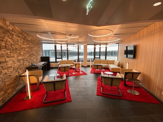 Geräumige, moderne Büro-Lounge mit bequemen, beigen Sesseln, Sofas, roten Teppichen, einer markanten Steinwand und stilvoller runder Pendelleuchte.
