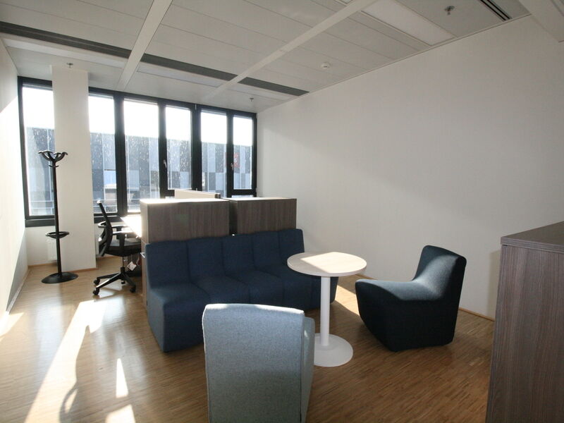 Heller moderner Büro-Lounge-Bereich mit blauem Sofa, Sesseln, einem kleinen runden Tisch und großen Fenstern mit Tageslicht