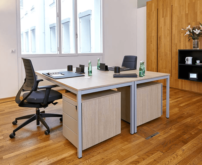 Bright modern office room with a double desk setup, ergonomic office chairs, wooden roll containers, and a large window.