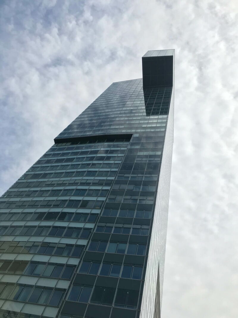 Low-angle view of a contemporary glass skyscraper with a distinctive top structure and cloudy sky background