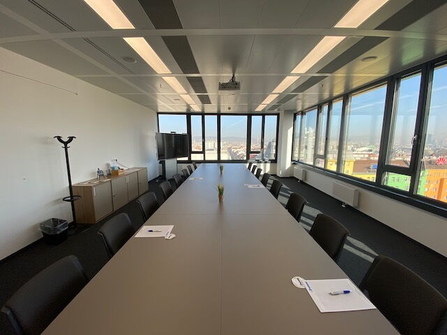 Ein geräumiger und moderner Konferenzraum mit einem langen Besprechungstisch, schwarzen ergonomischen Stühlen und großen, raumhohen Fenstern mit Panoramablick auf die Stadt.