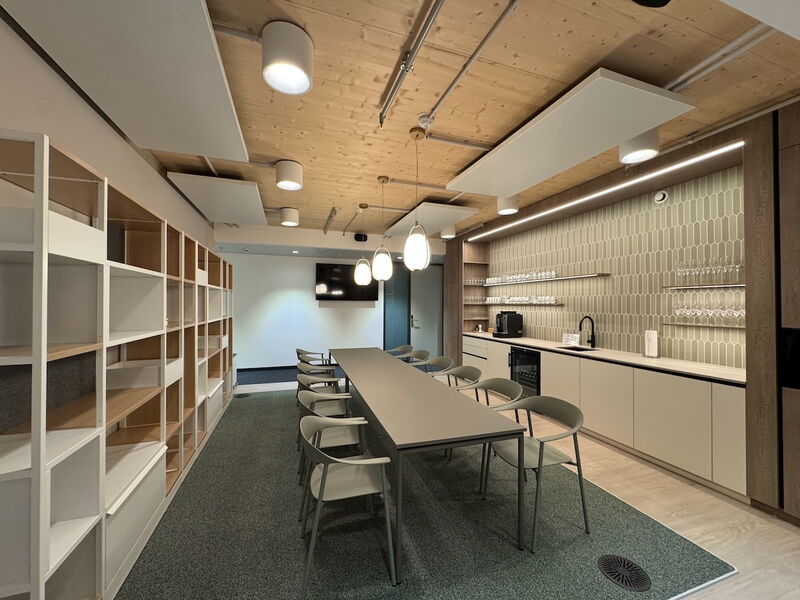 A modern office breakroom featuring a long conference table, kitchenette with a coffee machine, and wooden acoustic ceiling.