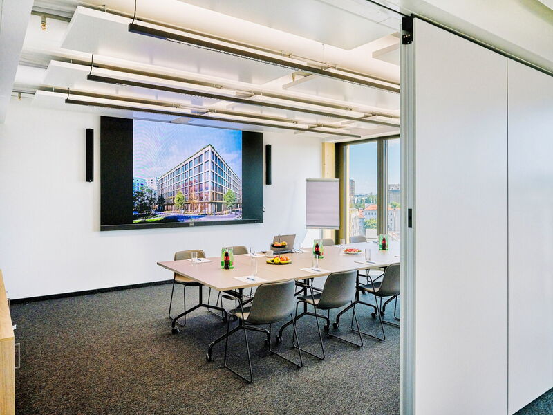 Bright and modern conference room featuring a large presentation screen displaying building architecture, a long meeting table with chairs, and drinks.