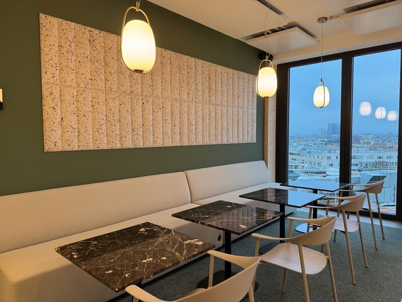 Modern office lounge interior featuring a long beige sofa, dark marble tables, pendant lights, acoustic wall panels, and a large window with a city view.