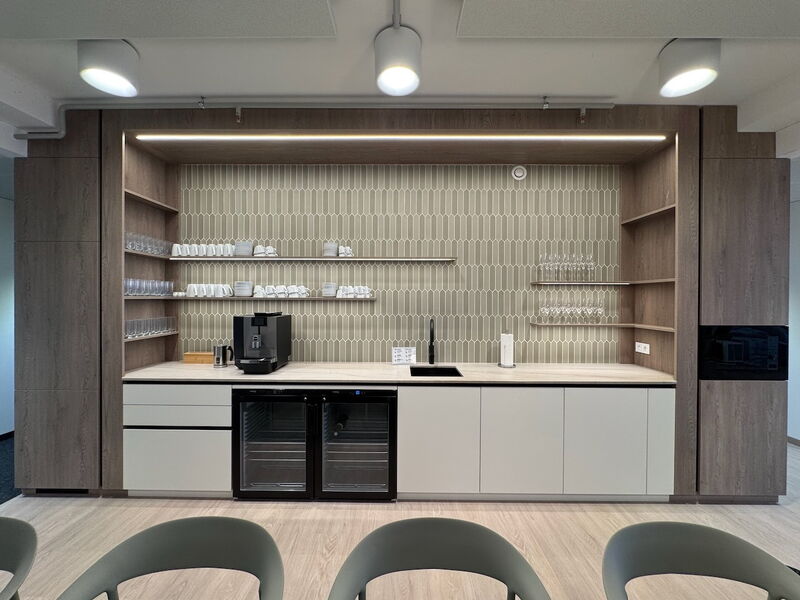 A sleek modern office kitchenette featuring wooden cabinetry, a coffee machine, a sink, glassware shelves, and a tiled backsplash.