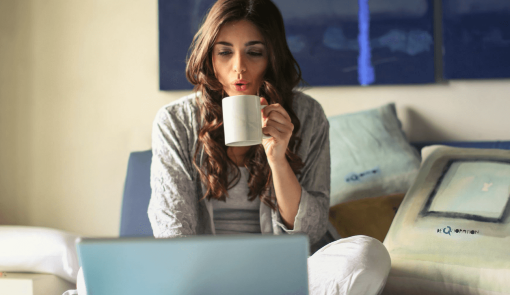 Junge Frau sitzt auf dem Sofa im Wohnzimmer, trinkt aus einer weißen Tasse und arbeitet am Laptop