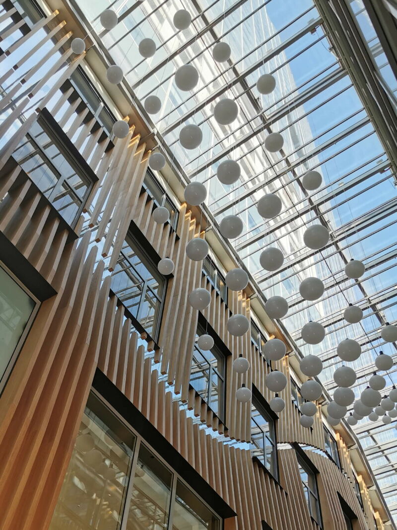 Looking up at a modern multi-story interior with a glass atrium roof and numerous hanging spherical light fixtures