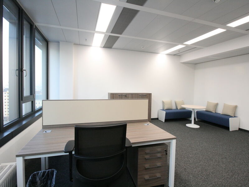 Modernes Büro mit Holztisch, Bürostuhl, großen Fenstern und einer Loungeecke mit blauen Bänken.