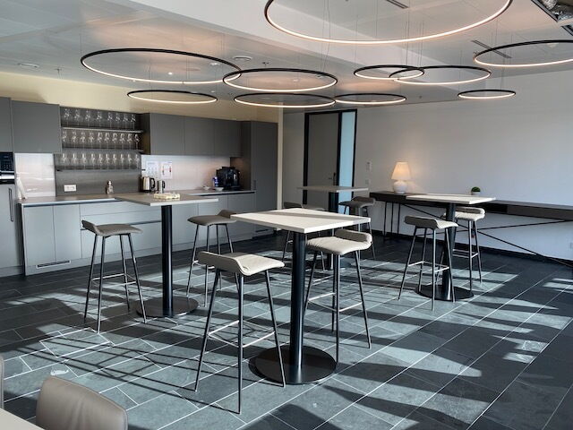 Modern office break area featuring high standing tables, bar stools, a kitchen with grey cabinetry, and unique ring lighting hanging from the ceiling.