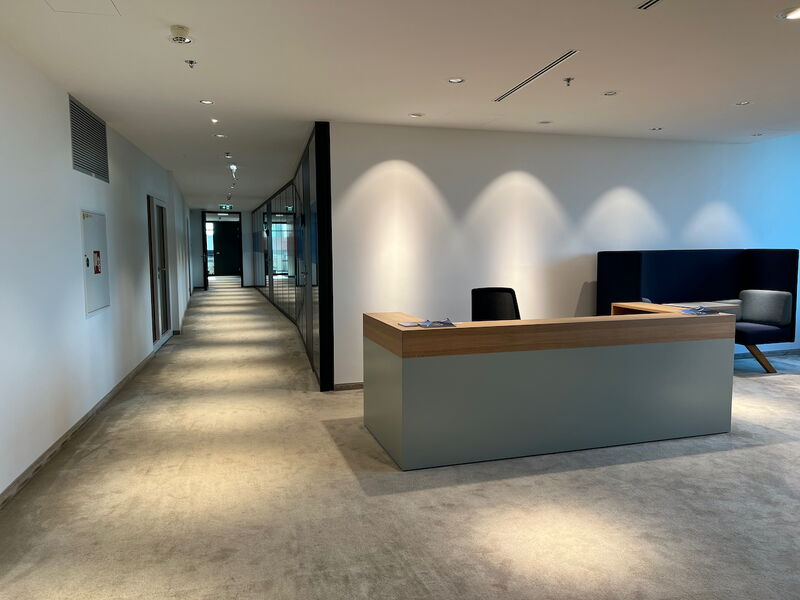 Modern office reception desk in a brightly lit hallway with a long carpeted corridor and glass partitioning