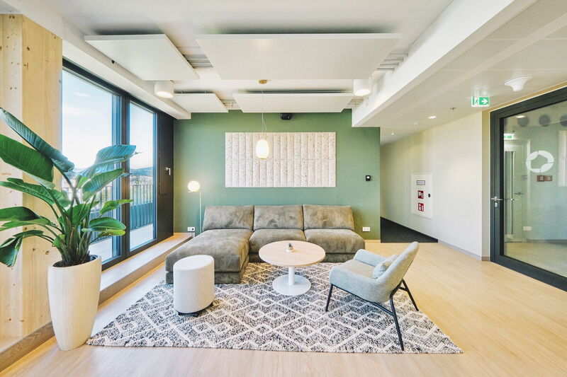 Modern office lounge area featuring a comfortable green sofa, acoustic ceiling panels, a green accent wall, and wooden flooring.