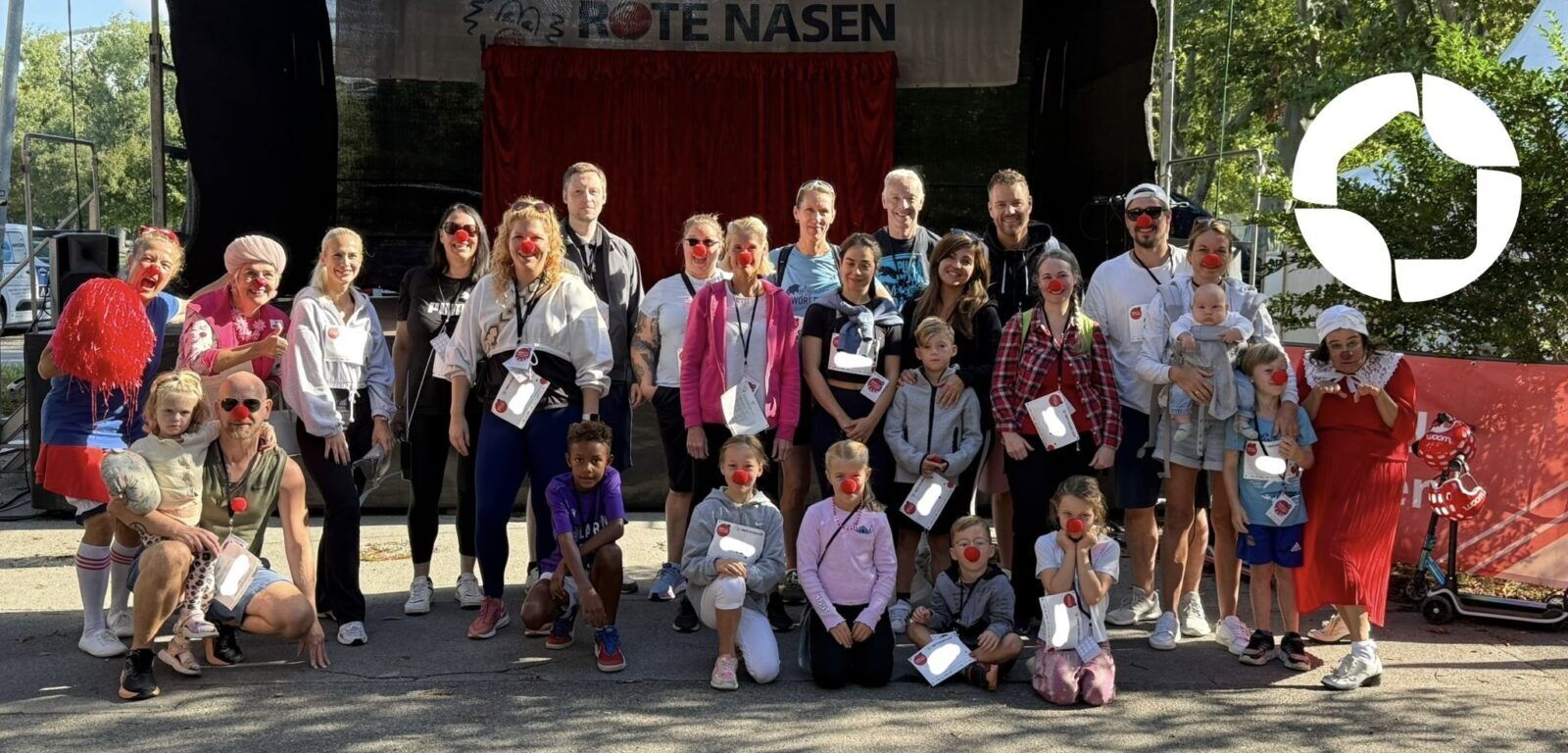 Eine große Gruppe von Erwachsenen und Kindern trägt rote Clownsnasen und posiert gemeinsam vor einer Bühne mit einem ROTE NASEN-Banner.