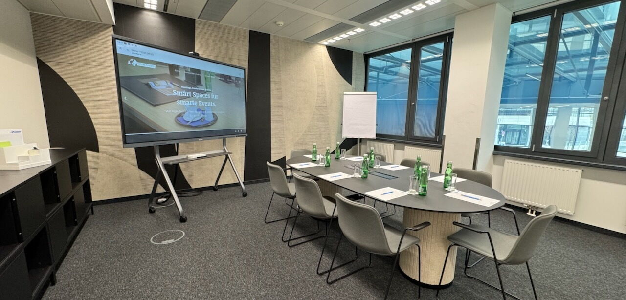 A modern, bright conference room featuring a large interactive digital screen on a stand, an oval meeting table with grey chairs, and large windows.