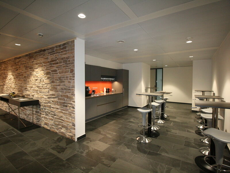 Moderne graue Büro-Teeküche mit orangener Rückwand, Einbau-Kaffeemaschine und Stehtischen mit weißen Hockern.