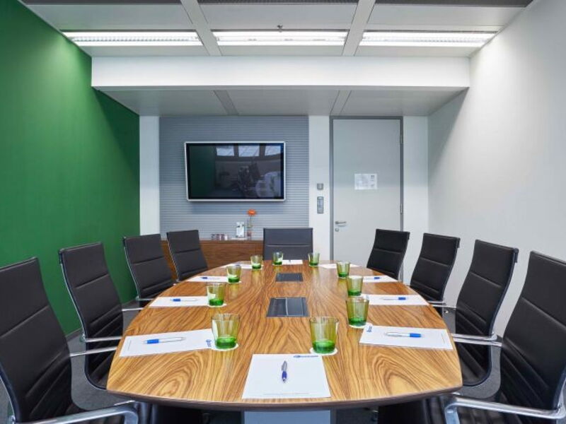 Spacious and modern conference room featuring a large oval wooden table, black ergonomic chairs, and a green accent wall.