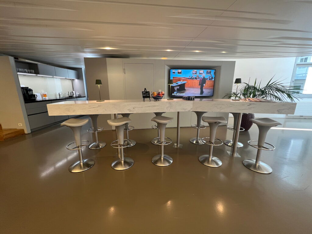 Modern office breakroom featuring a long marble-top bar counter, gray bar stools, a kitchen area, and a large TV screen on the wall.