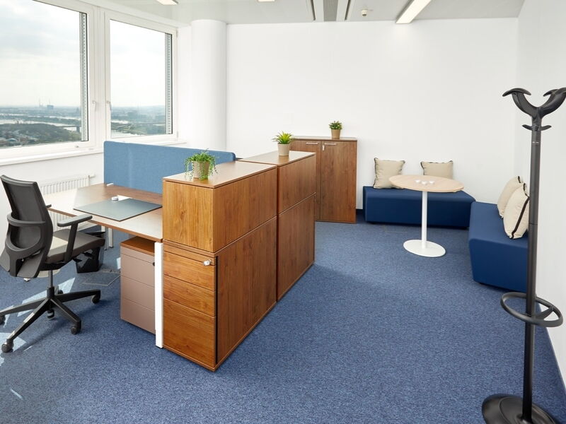 Helles, modernes Büro mit Holztischen, blauen Sichtschutzwänden, Zimmerpflanzen und großen Fenstern mit Blick auf die Stadt.