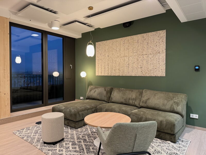 Cozy office lounge interior featuring a large green corduroy sectional sofa, a wooden coffee table, and sound-absorbing panels on a green wall.