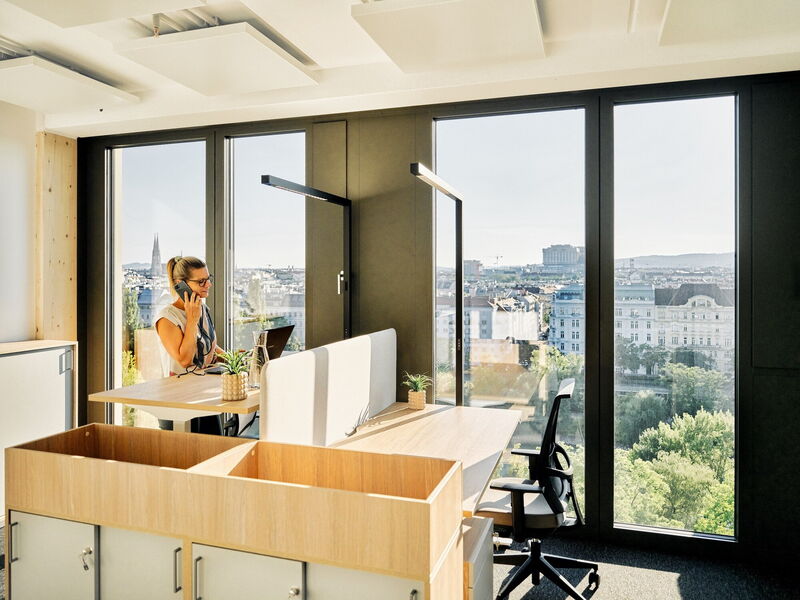 Businesswoman making a phone call at an ergonomic standing desk in a modern office with large panoramic windows overlooking the city skyline