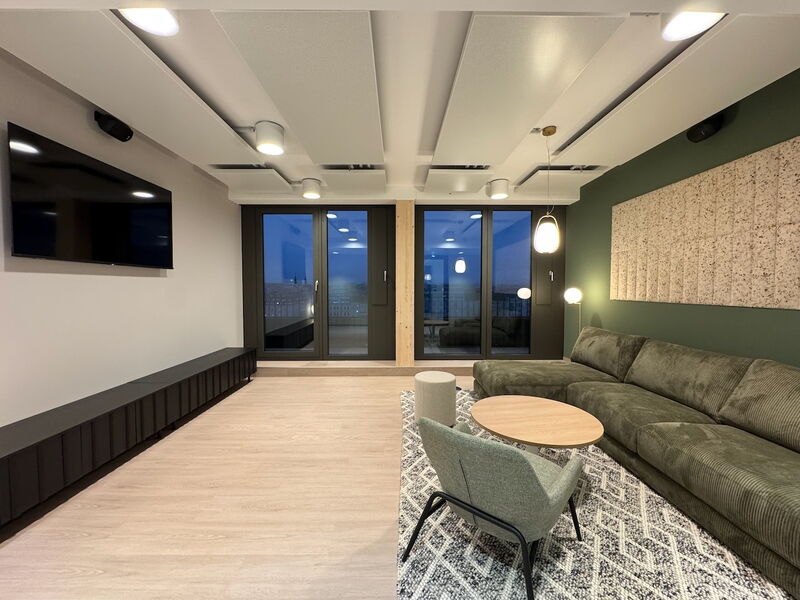 Spacious modern office lounge area featuring a large green corduroy sofa, acoustic ceiling and wall panels, a TV screen, and large windows at dusk.