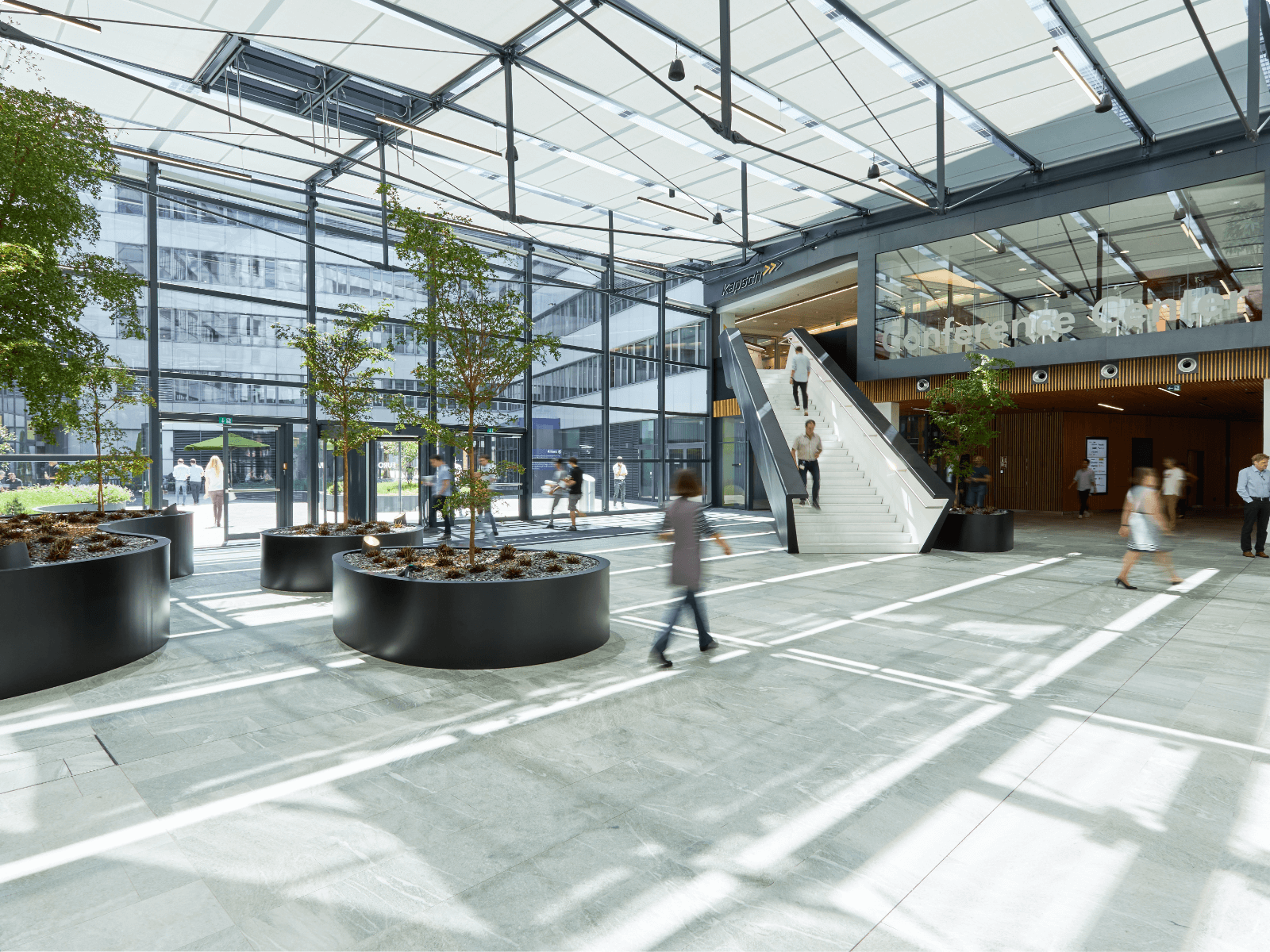 Bright modern office building atrium featuring large glass windows, indoor trees in planters, and a grand staircase leading to a conference center.