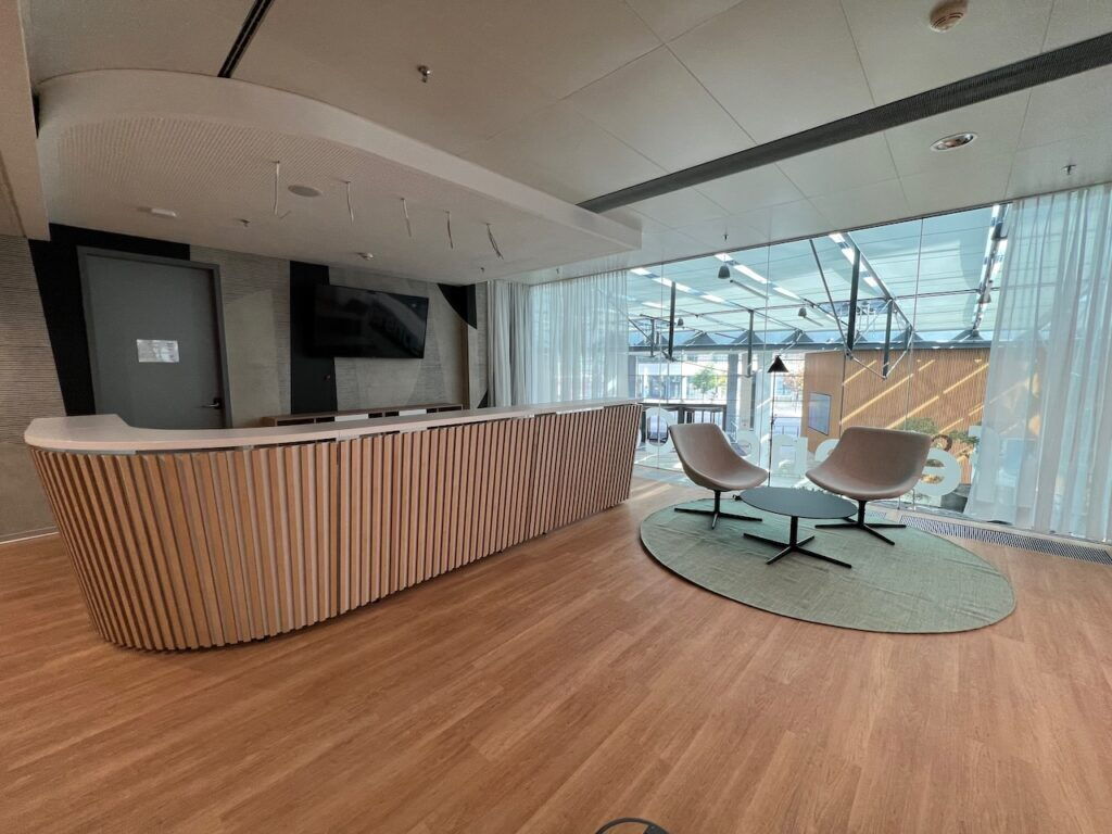 Modern office reception area featuring a large curved wooden counter, comfortable armchairs on a green rug, and large glass windows with natural light.