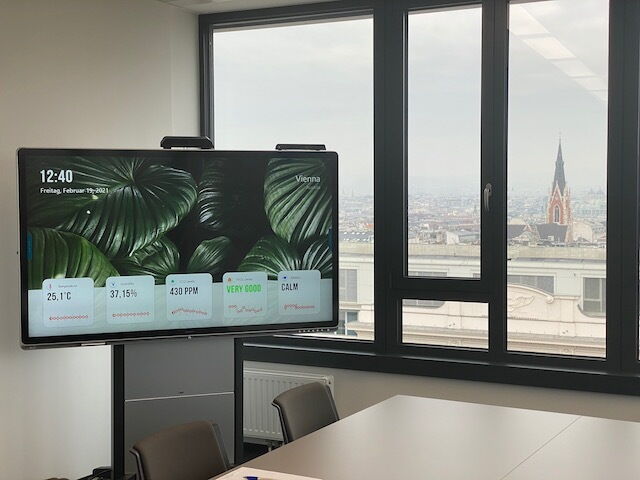 Ein moderner Konferenzraum mit einem großen interaktiven Bildschirm, der Raumklimadaten anzeigt, und Blick auf Wien aus dem Fenster.