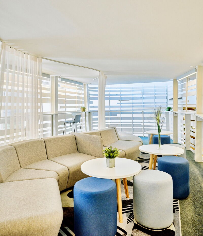 Geräumiger, moderner Loungebereich mit einem großen, geschwungenen beigen Sofa, kleinen weißen Beistelltischen, blauen und grauen Hockern sowie großen Fenstern mit weißen Vorhängen.