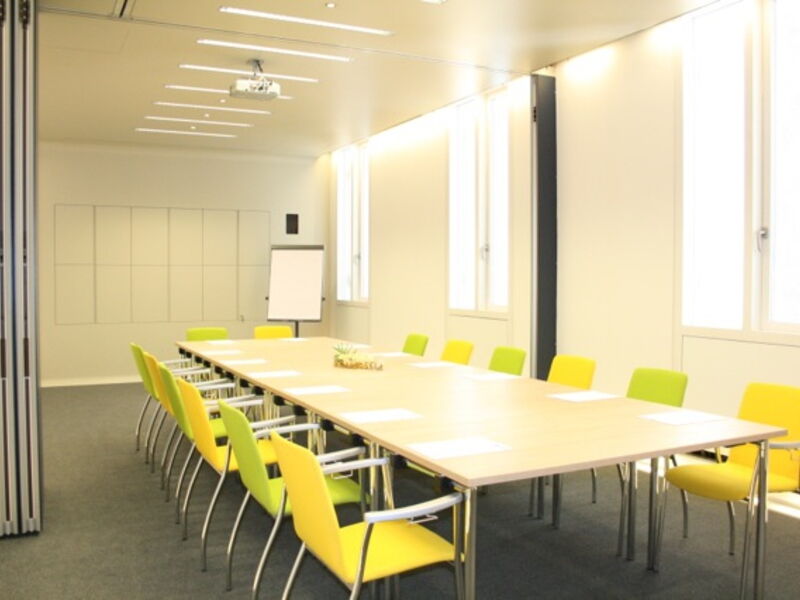 Heller und geräumiger moderner Konferenzraum mit einem langen Holztisch, umgeben von hellgrünen und gelben Stühlen, einem Beamer und großen Fenstern.