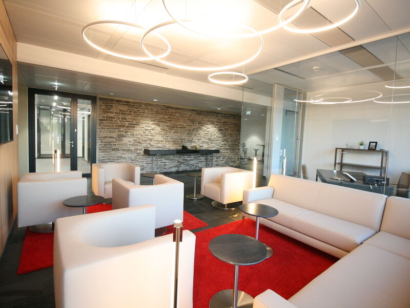Zeitgenössischer Büro-Lounge-Bereich mit modernen weißen Sesseln, Sofas, runden Leuchten und einem auffälligen roten Teppich auf Steinboden