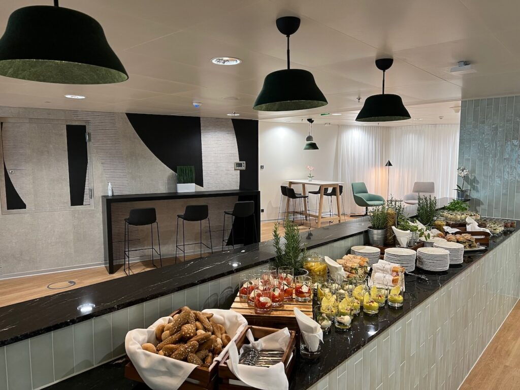 A modern office canteen and break room featuring a long buffet counter with fresh food, pastries, and appetizers, alongside bar seating and green pendant lights.