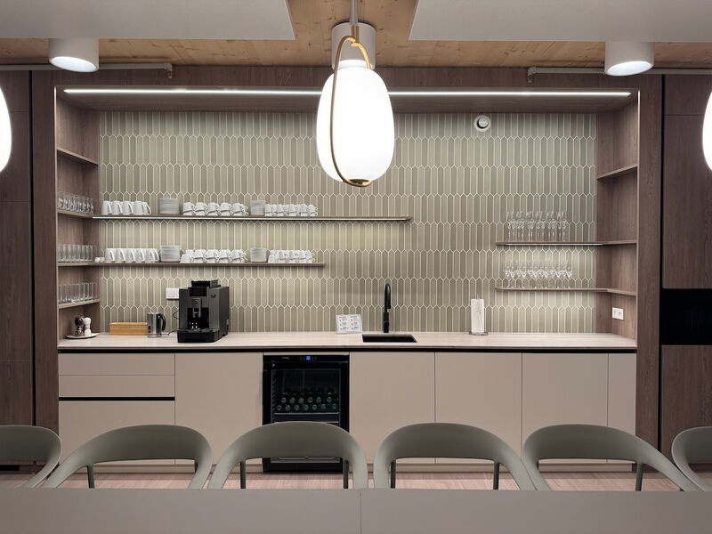 Sleek office breakroom kitchen featuring beige picket wall tiles, wooden shelving, built-in espresso machine, wine cooler, and modern pendant lighting.