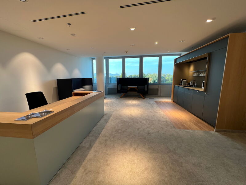 Spacious modern office space featuring a reception desk in the foreground, a built-in kitchenette on the right, and comfortable seating areas by the large windows.