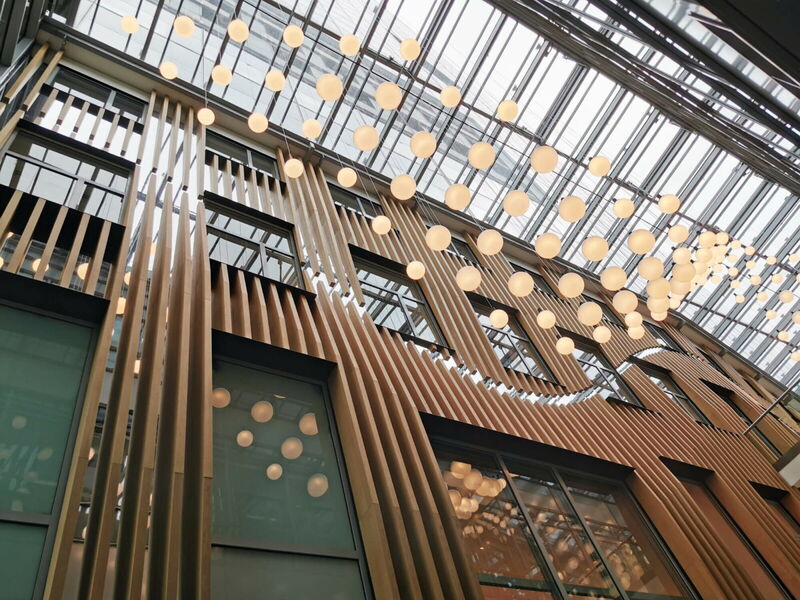 Interior view of a modern building featuring vertical wooden lamellas, large glass windows, and a dramatic glass ceiling with hanging spherical lights.