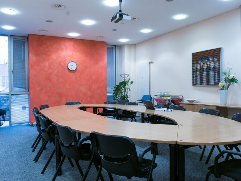 Ein großer hölzerner, U-förmiger Konferenztisch umgeben von schwarzen Bürostühlen in einem hellen Besprechungsraum mit roter Akzentwand