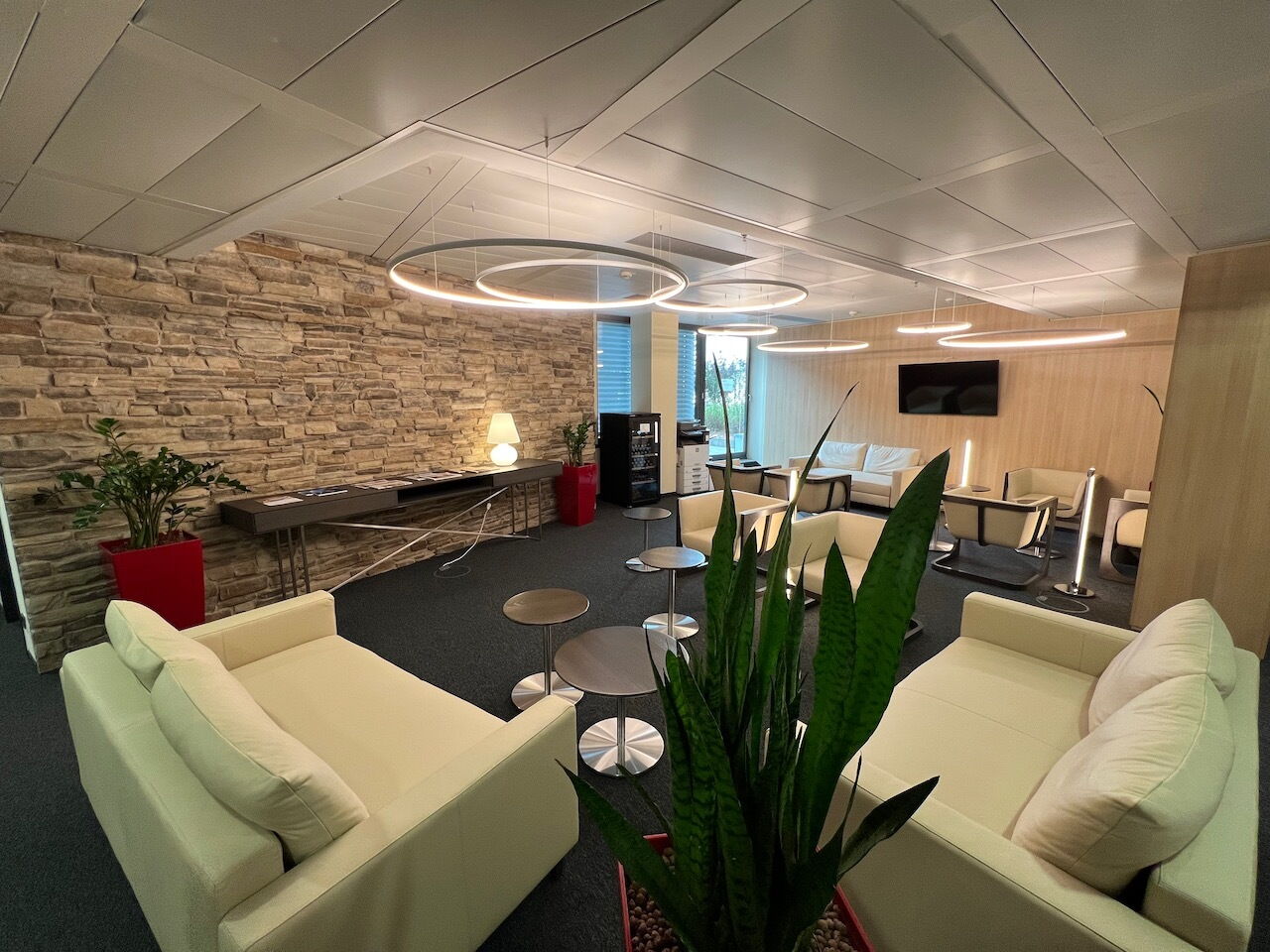 Spacious modern office lounge with comfortable white sofas, a rustic stone accent wall, and circular pendant lights.