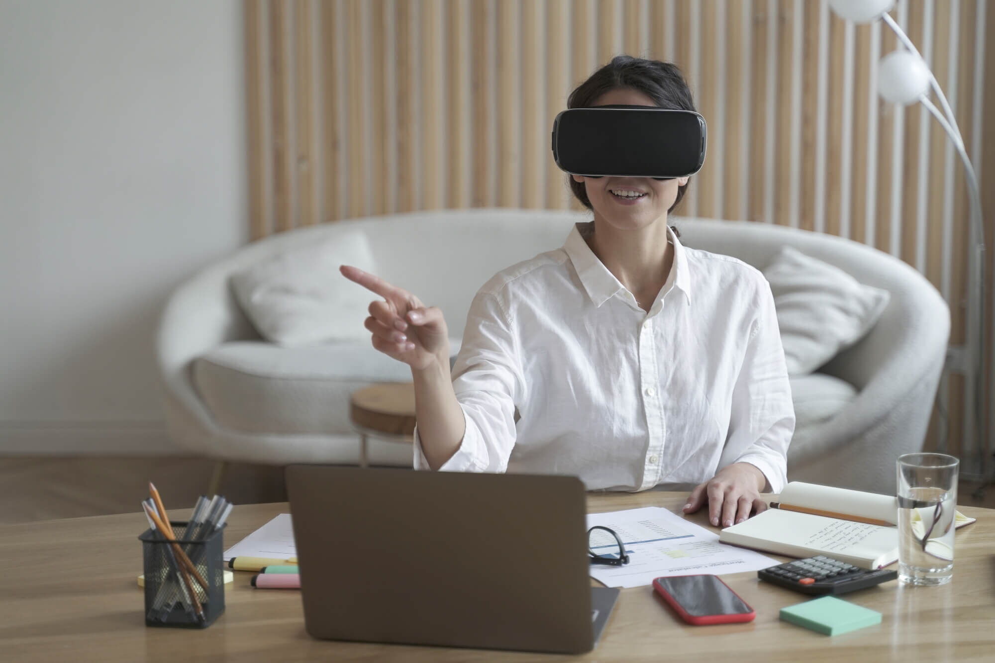 Smiling woman wearing VR goggles pointing at virtual screen while sitting at office desk with laptop