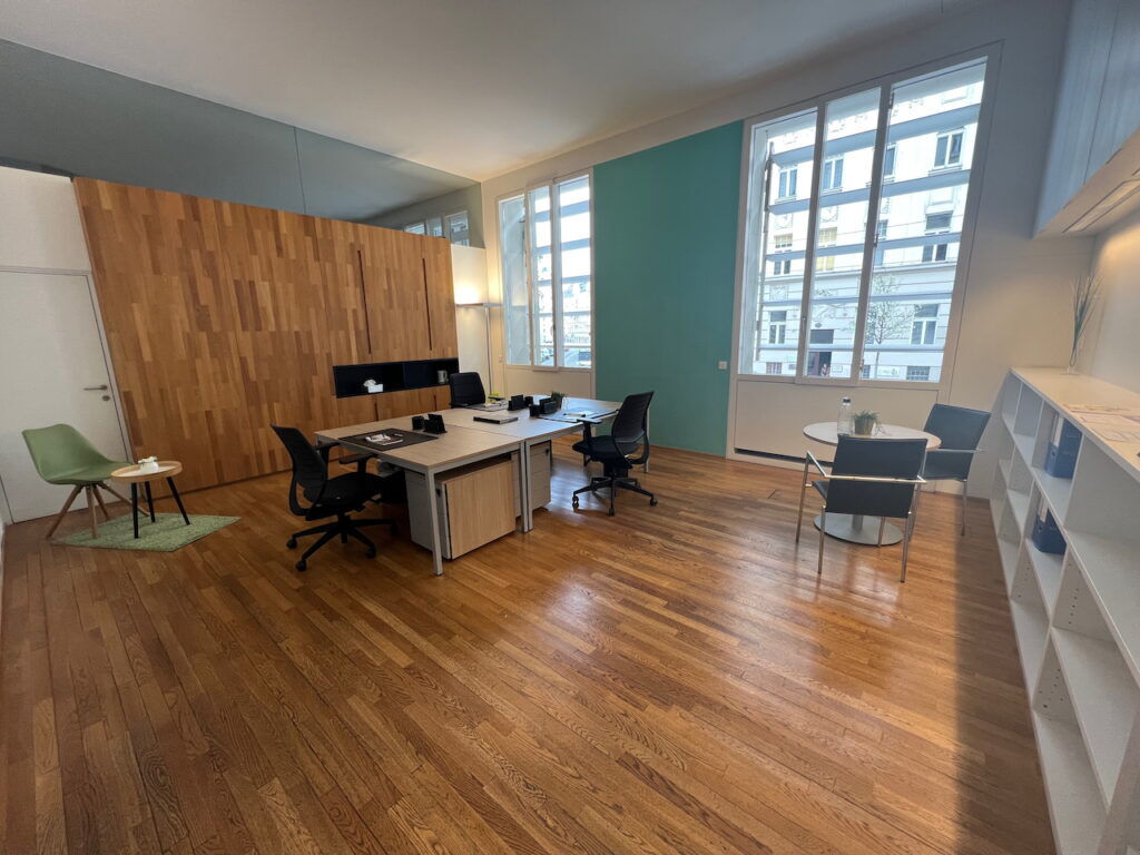 Bright and spacious modern office with wooden flooring, desks, ergonomic chairs, and large windows overlooking city buildings.