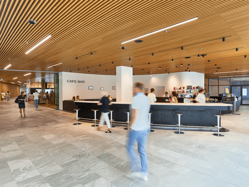 Modernes Galerie-Interieur mit einer schicken Café-Bar, Holzlamellendecke und Besuchern auf Steinboden