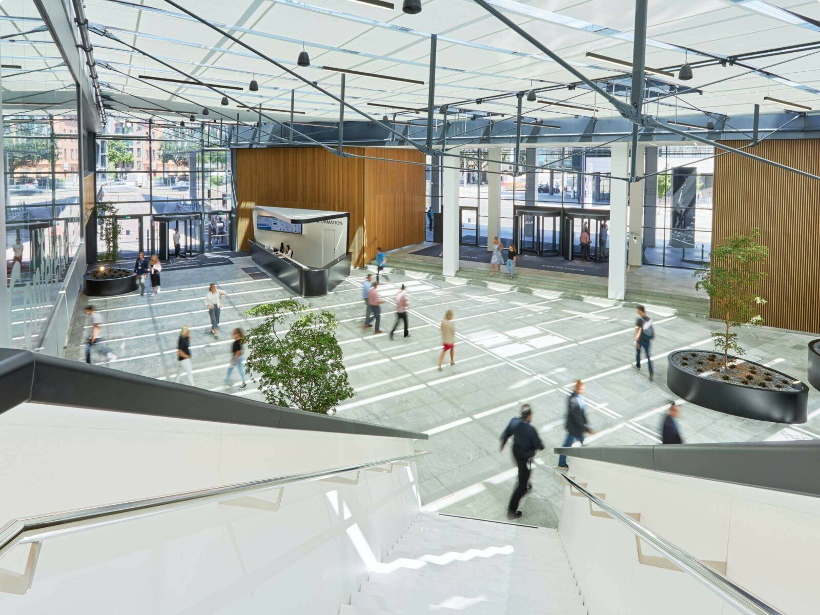 Spacious modern office lobby with a high glass roof, wooden accents, and people walking across the floor, viewed from above.
