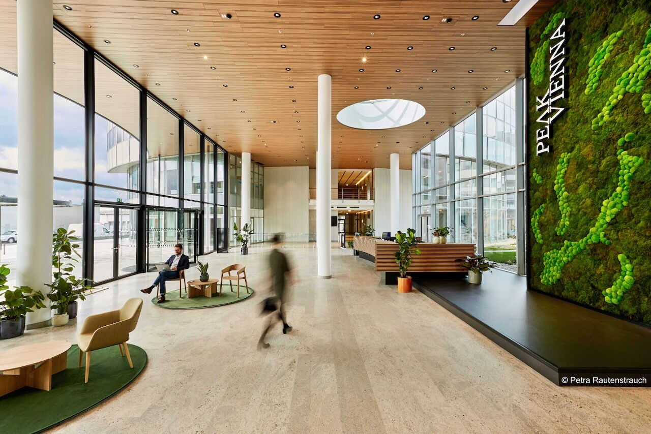 Geräumiges und modernes Foyer eines Bürogebäudes im Peak Vienna mit großen Glasfronten, Holzdecke und einer auffälligen grünen Mooswand mit Firmenlogo