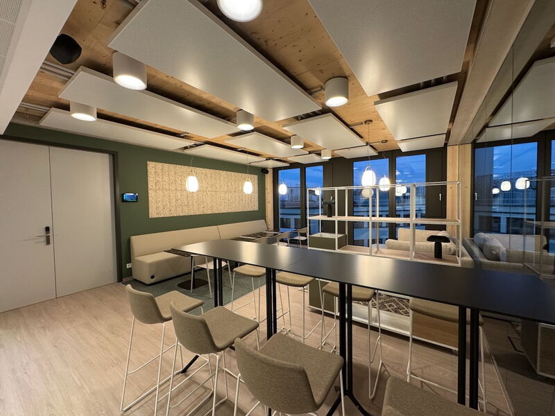 Spacious modern office breakout area featuring a long black high table, bar stools, comfortable sofas, and suspended acoustic ceiling panels.